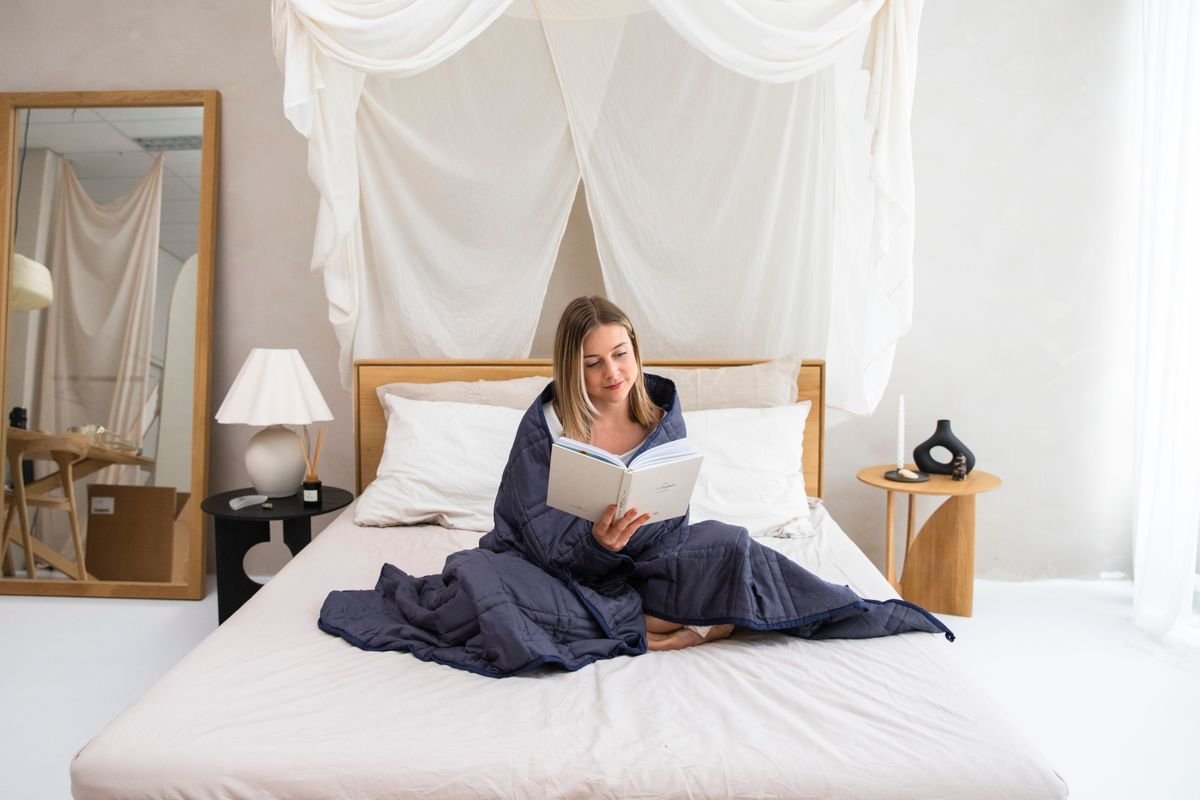 Mulher sentada na cama enrolada em um cobertor ponderado azul lendo um livro, primeira forma de usar o cobertor ponderado
